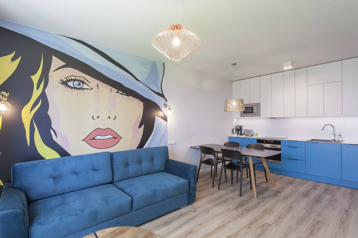 Un salon moderne avec un canapé bleu, un coin repas comprenant une table et des chaises, et une cuisine avec des meubles blancs et un plan de travail bleu.