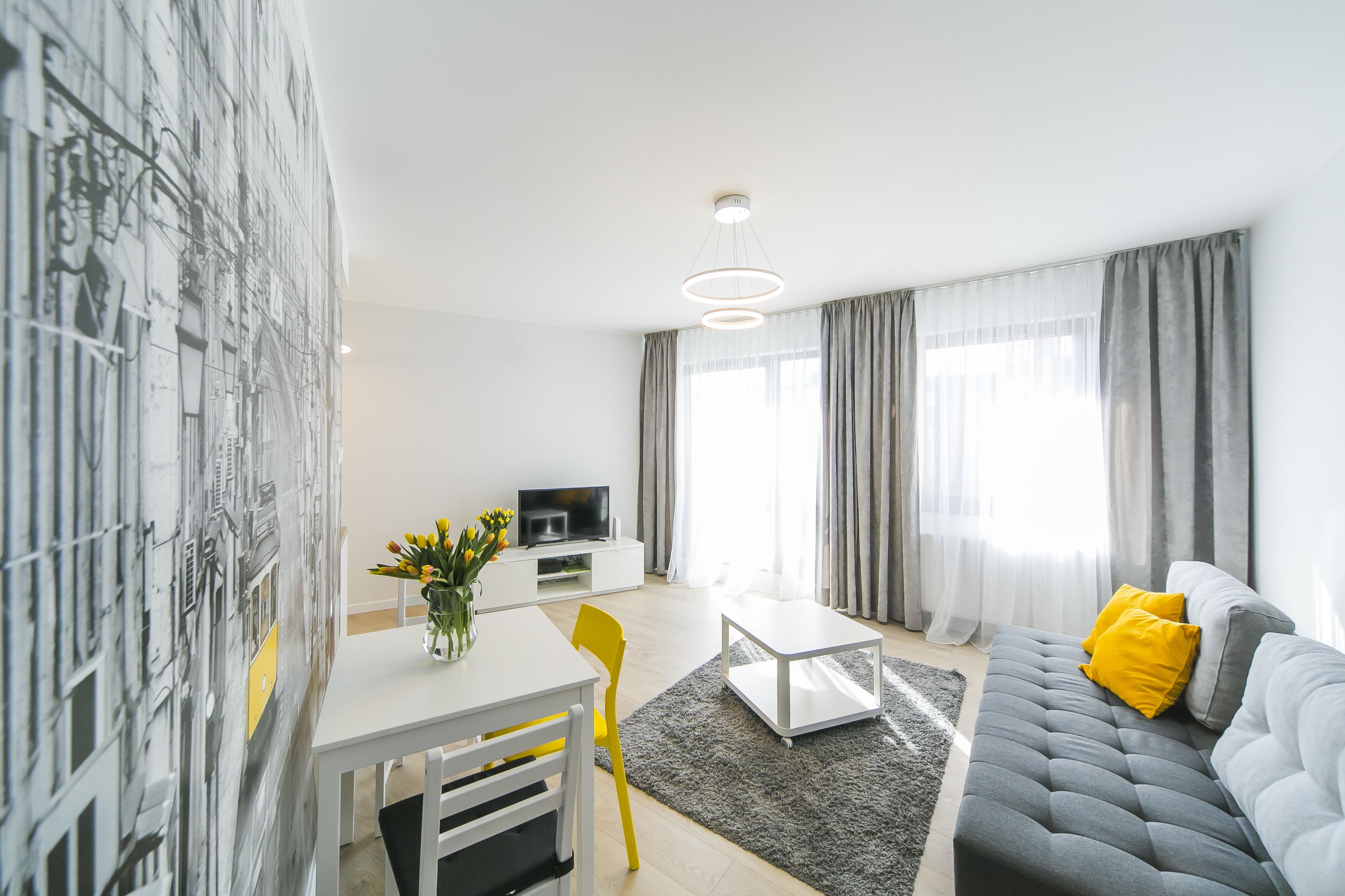 A modern living room with a grey sofa, a white coffee table, and a yellow chair. There is a dining table with a vase of flowers and a flat-screen TV on a white cabinet.