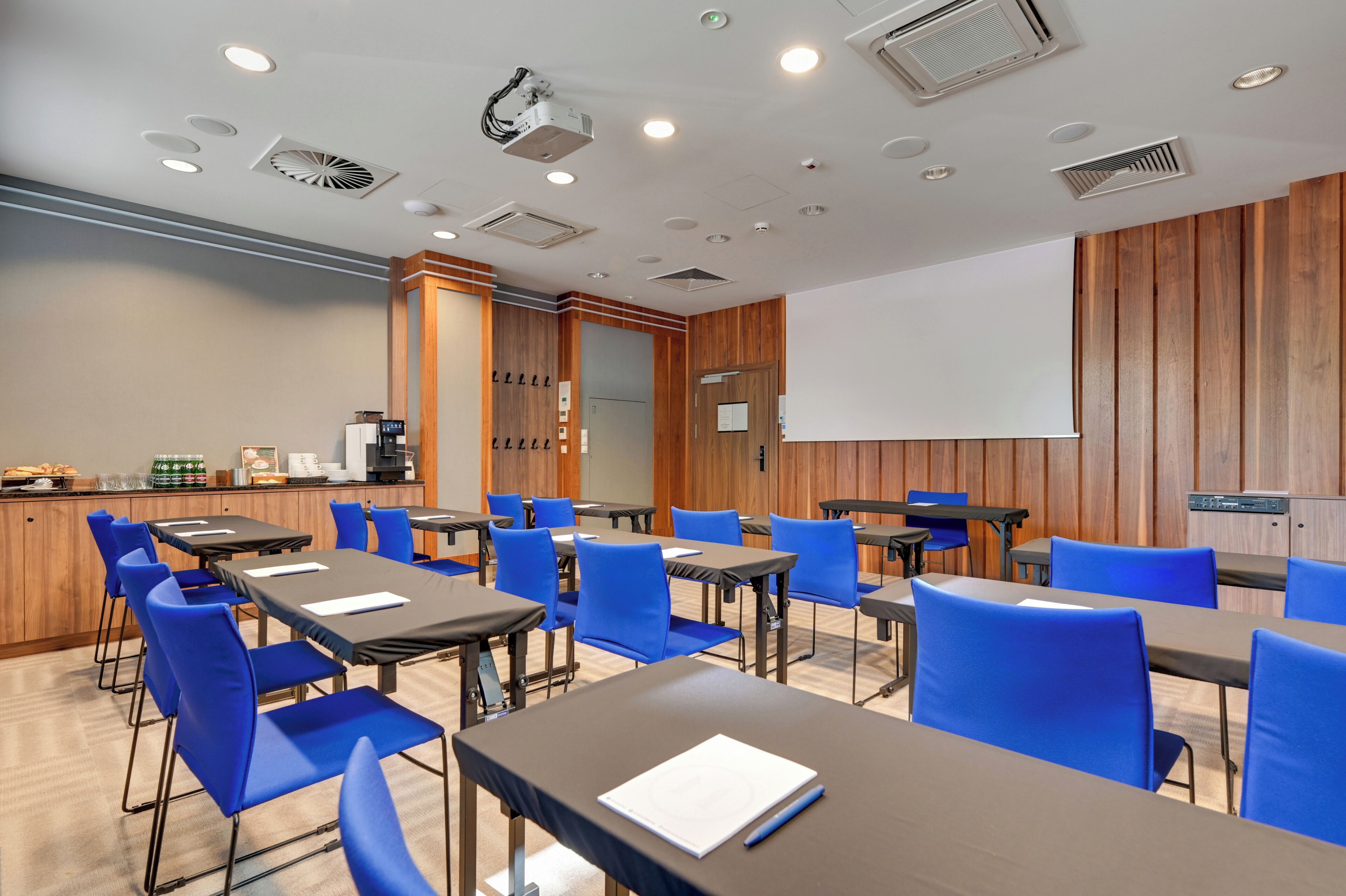 A conference room with tables, chairs, a projector screen, and a coffee station.