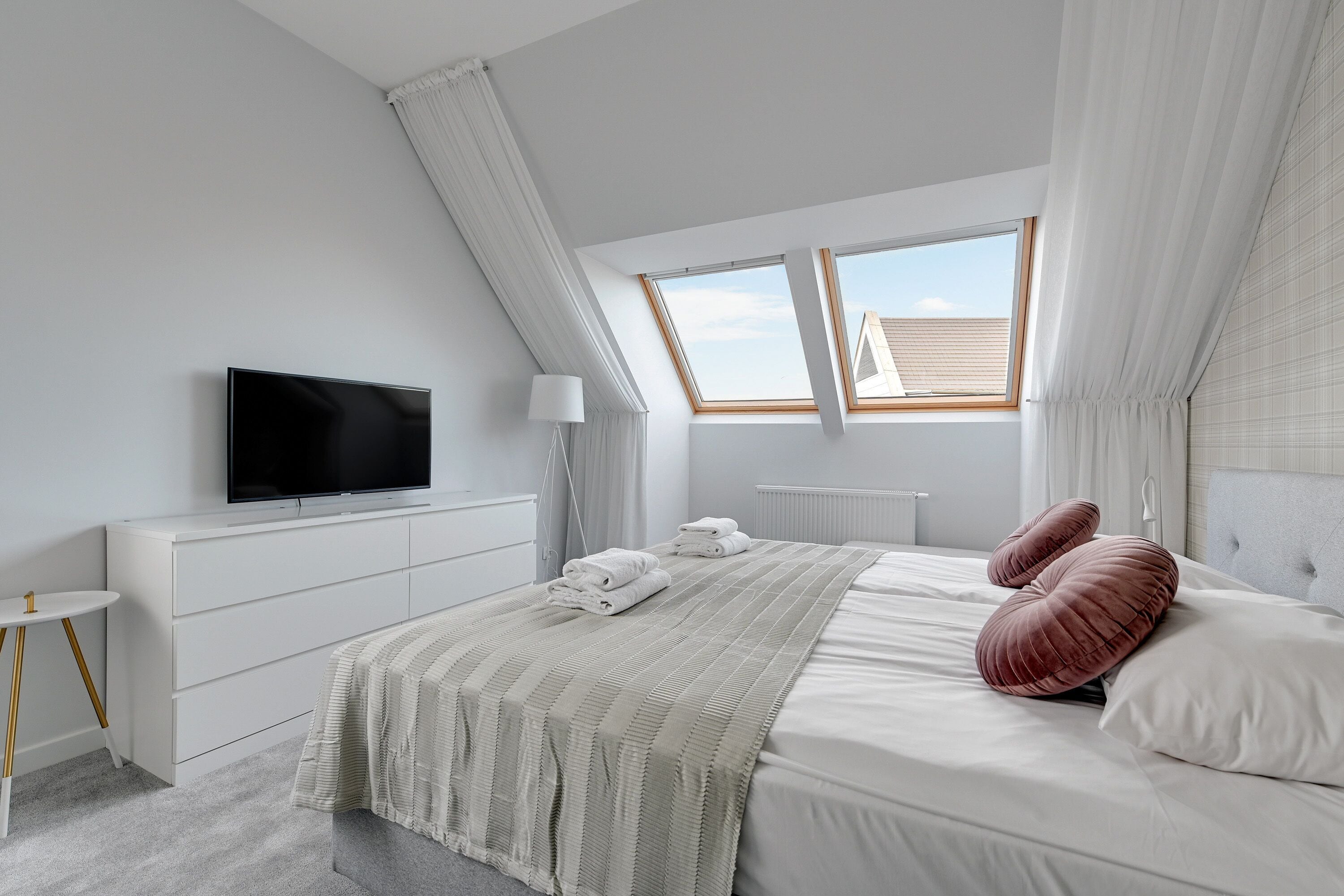 A modern bedroom with a flat-screen TV, a neatly made bed, a bedside table, and a window with curtains.