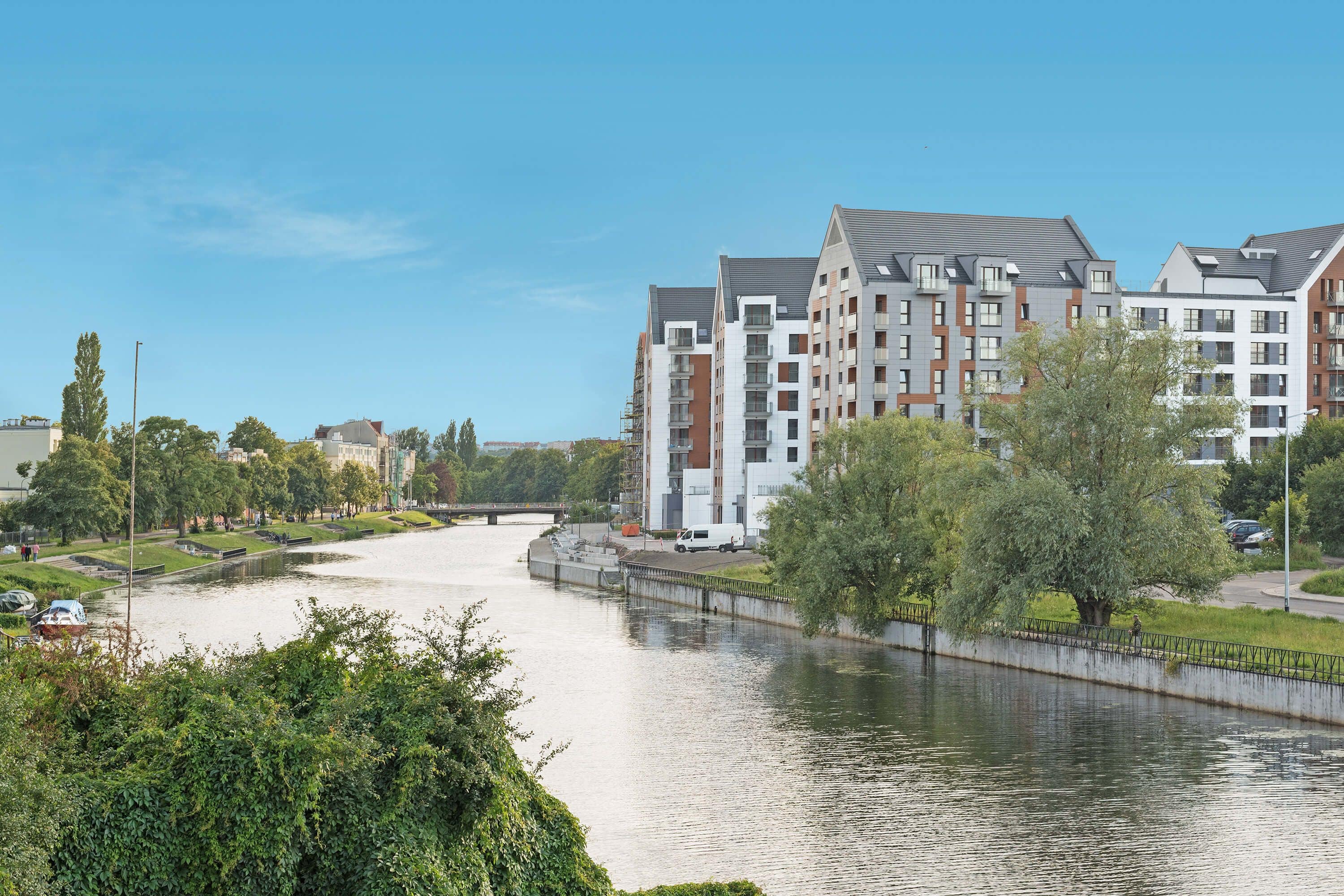 A riverside view of a modern apartment complex with multiple buildings, a bridge, and lush greenery.