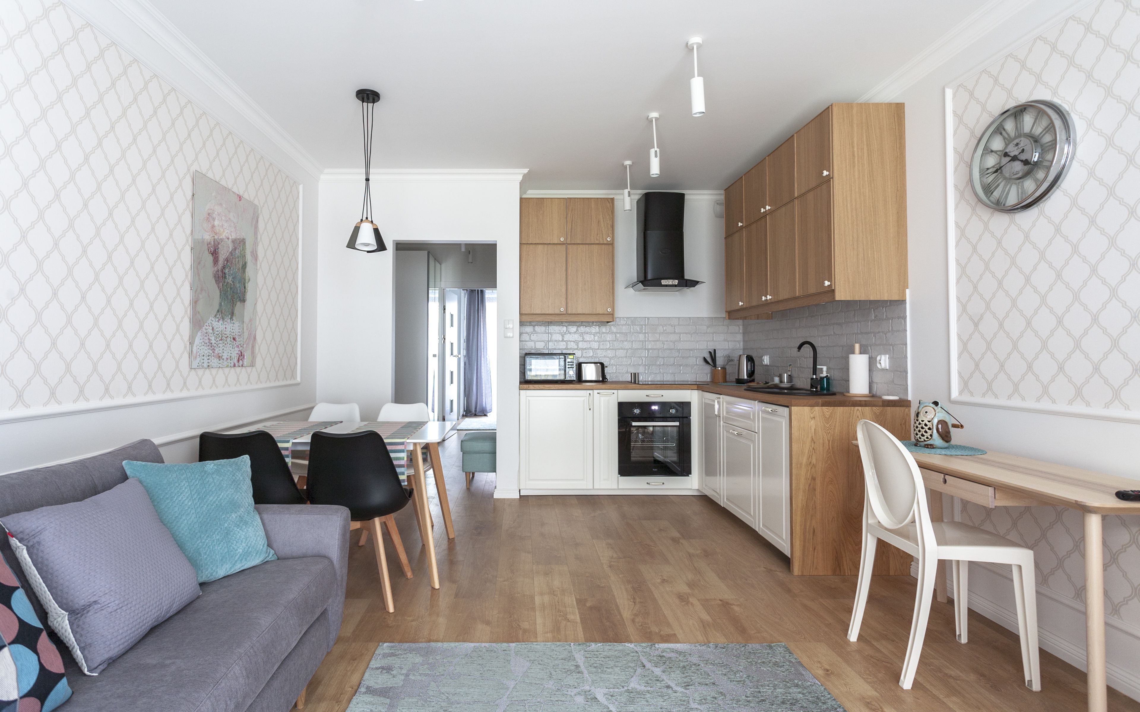 A modern kitchen with wooden cabinets, a built-in oven, and a dining area with a grey sofa and white chairs.