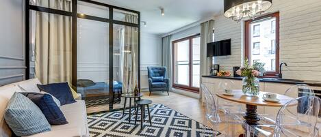 A modern living room with a sofa, a patterned rug, a dining table with clear chairs, and a chandelier.