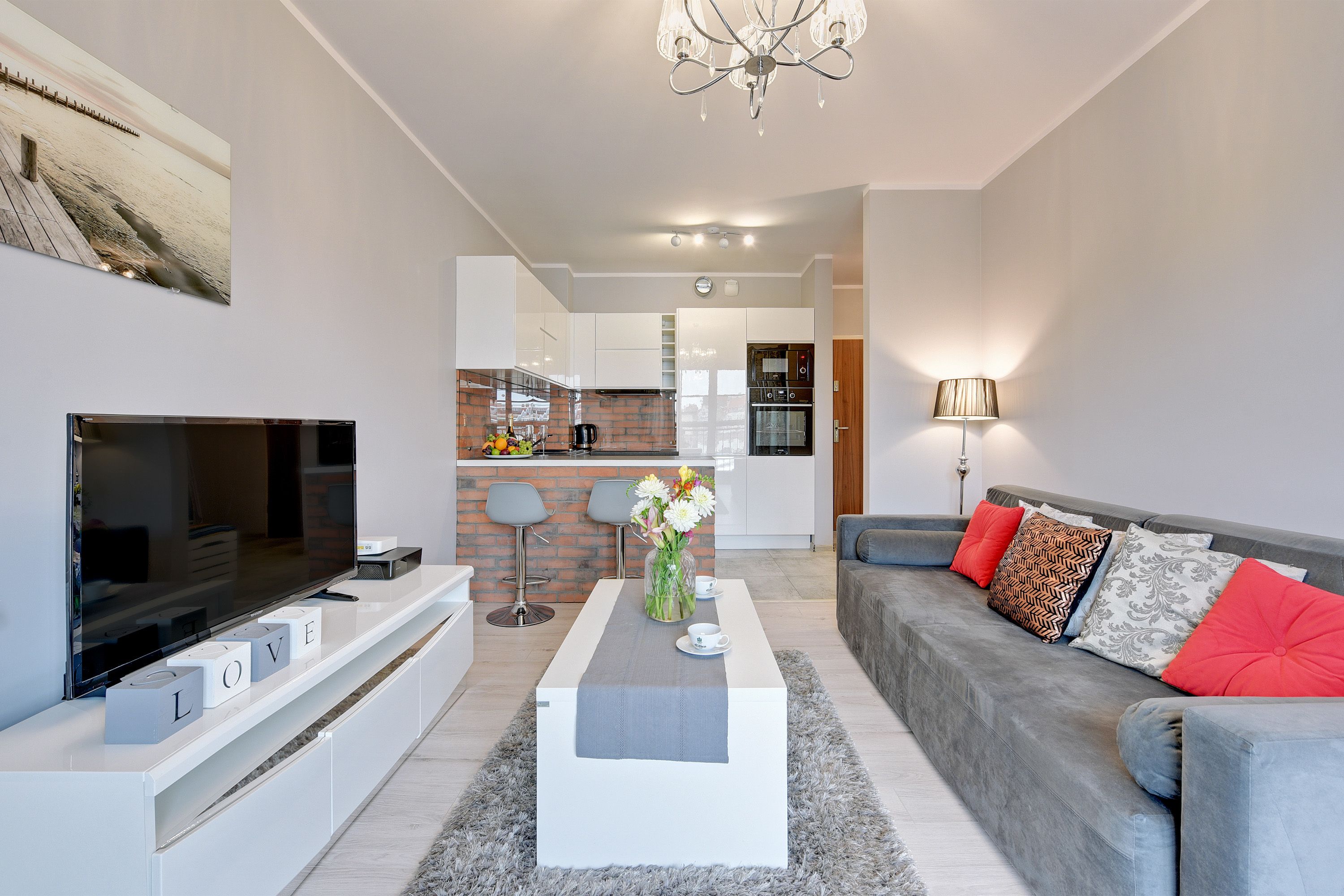 A modern living room with a flat-screen TV, a coffee table with a vase of flowers, and a grey sofa with red cushions.