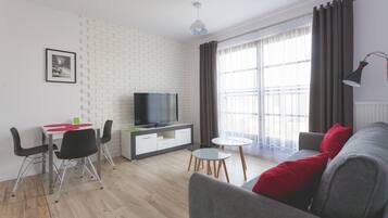A modern living room with a flat-screen TV, a grey sofa, a small round table, and a dining area with a white table and black chairs.