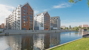 A modern apartment building with multiple balconies, a river in front, and a grassy area. - GRANO APARTMENTS Gdańsk Motława SPA & Wellness (Gdansk)