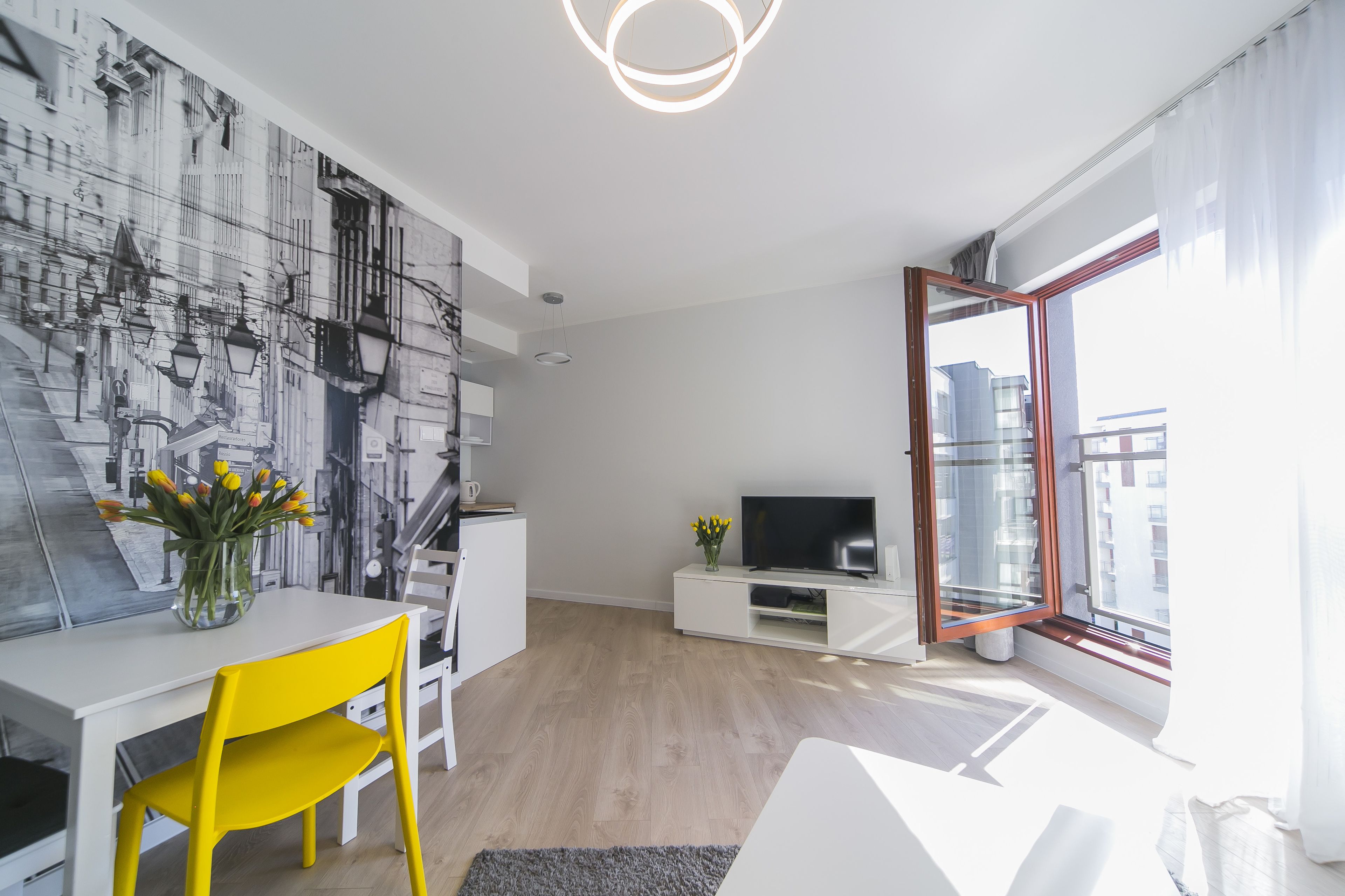 Una sala de estar moderna con comedor, un televisor sobre un mueble blanco y un ventanal con cortinas.