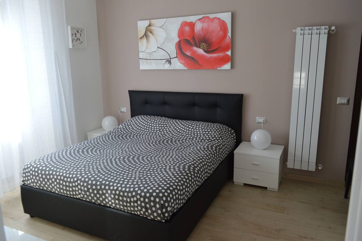 Een slaapkamer met een bed, nachtkastjes, een radiator en een aan de muur bevestigd kunstwerk.