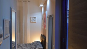 A narrow hallway leading to a bedroom with a bed, bedside table, and a lamp.