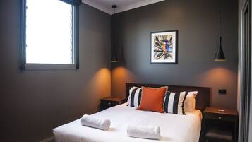 A neatly made bed with white linens and two rolled towels, a framed artwork on the wall, and two bedside lamps.