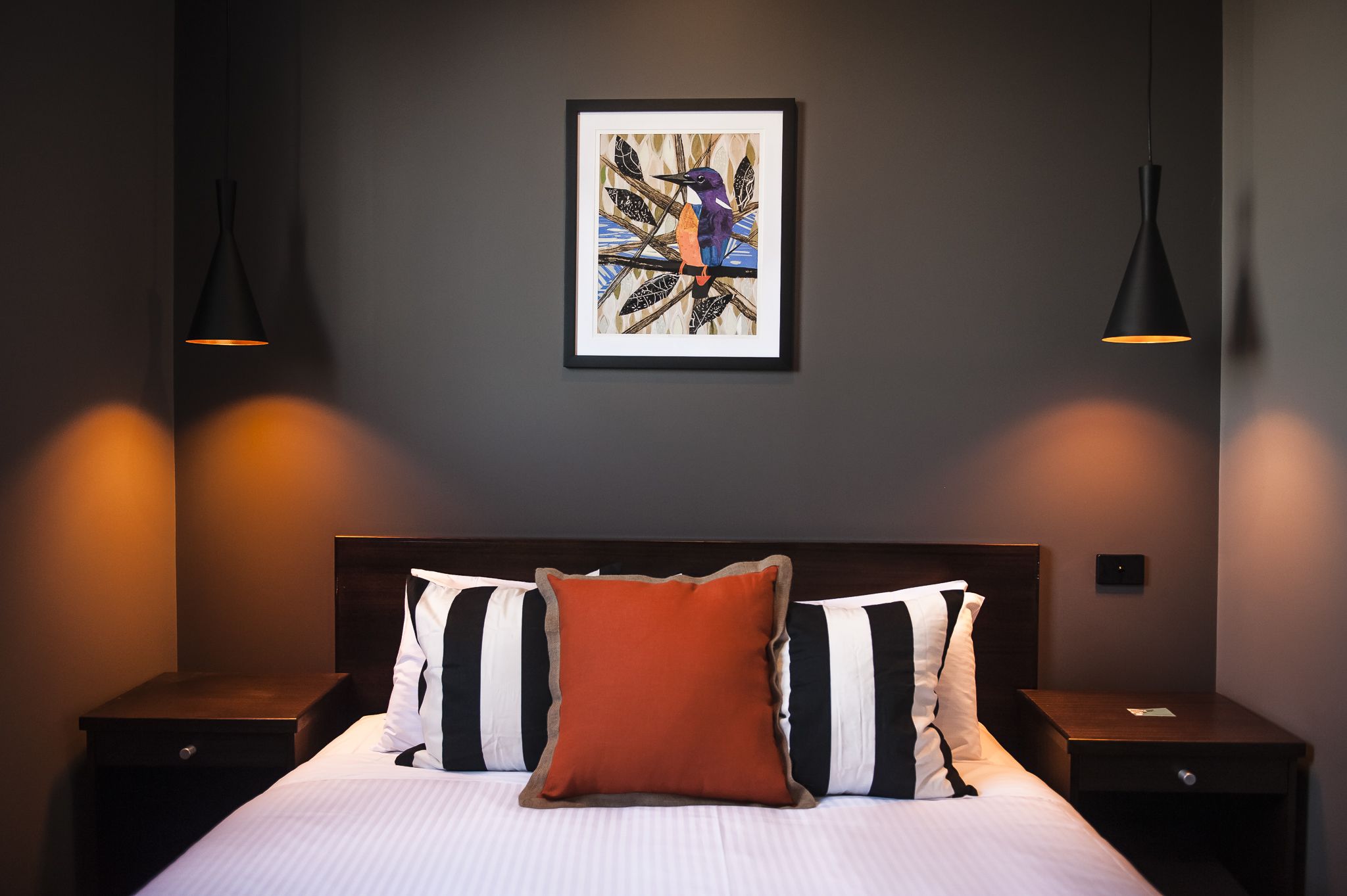 Una cama bien hecha con almohadas a rayas, un cuadro abstracto enmarcado en la pared y dos lámparas de noche.
