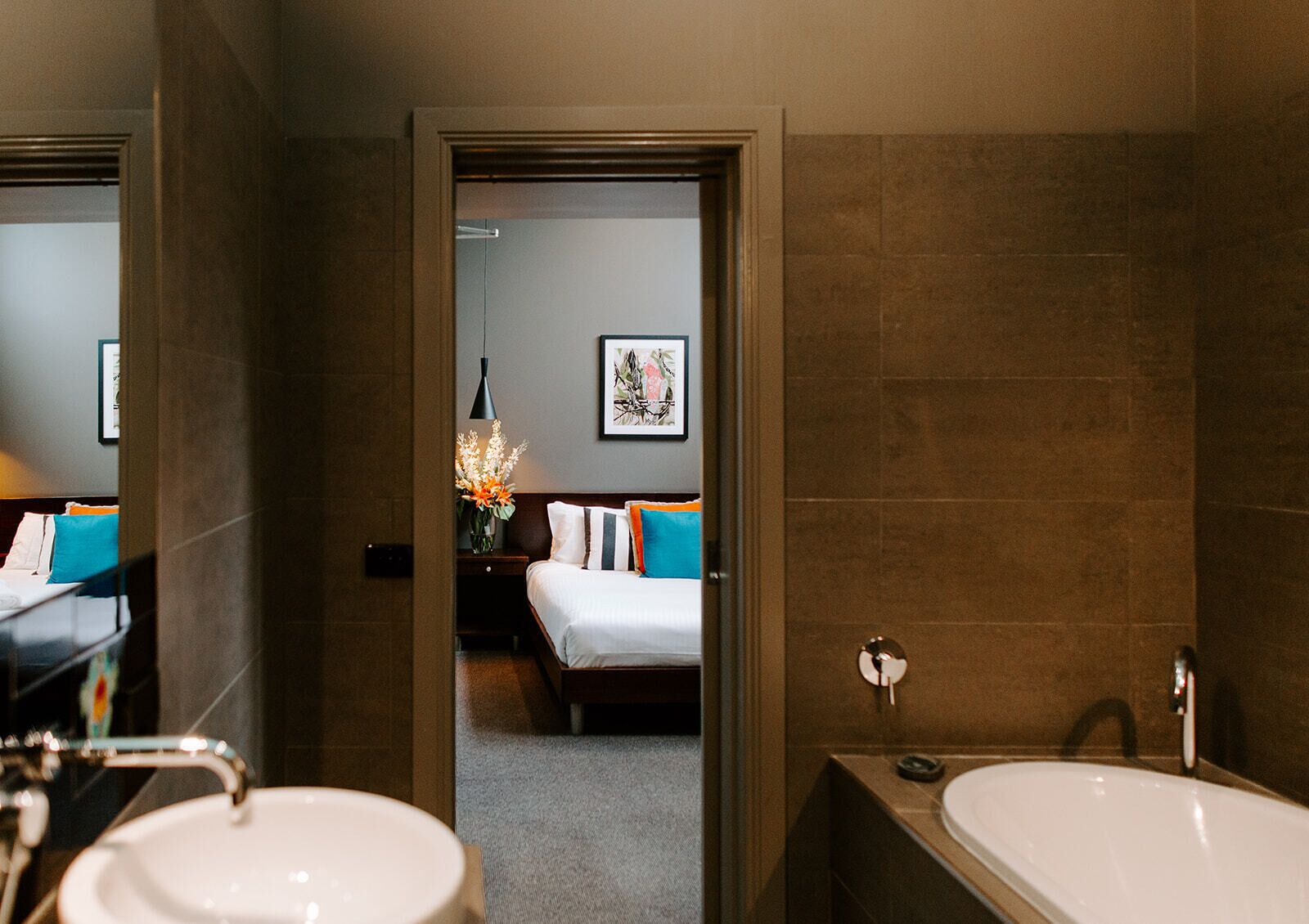 A hotel bathroom with a large bathtub, a sink, and a mirror. There is a bed visible through an open door in the background.