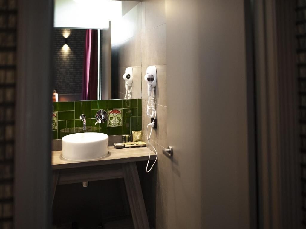 A bathroom with a white sink, green tiles, a mirror, and two hair dryers mounted on the wall.