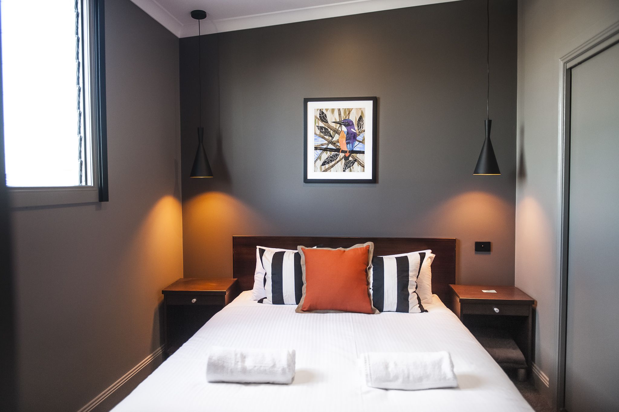 A neatly made bed with white linens and two towels, a framed abstract painting on the wall, and two bedside lamps.