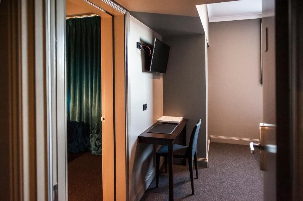 A compact hotel room with a single bed, a small desk with a chair, a television mounted on the wall, and a door leading to another room.