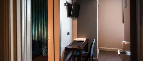 A compact hotel room with a single bed, a small desk with a chair, a television mounted on the wall, and a door leading to another room.