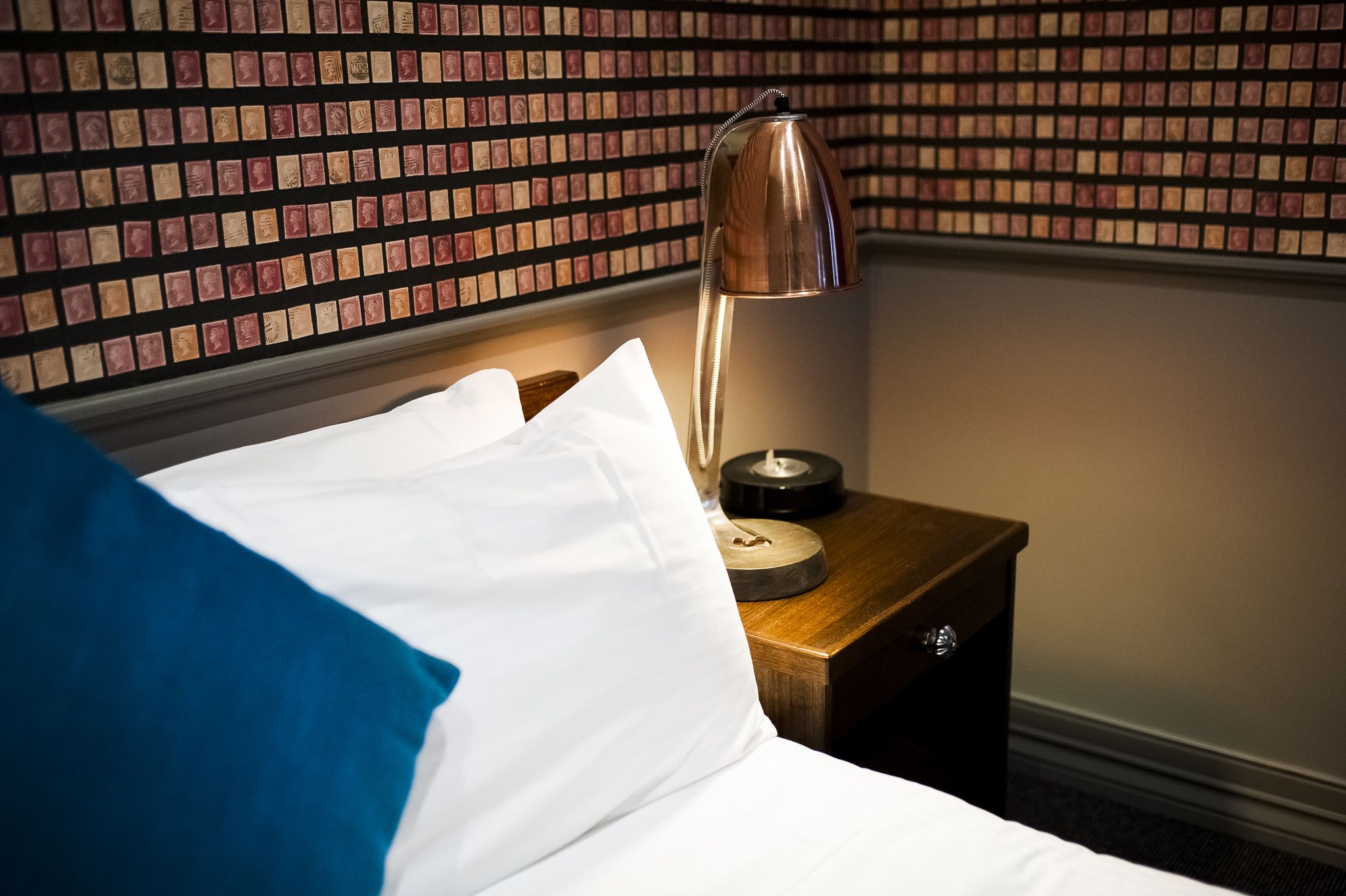 Una cama bien hecha con almohadas blancas y azules, una mesita de noche de madera con lámpara y una pared con diseño de baldosas estampadas.