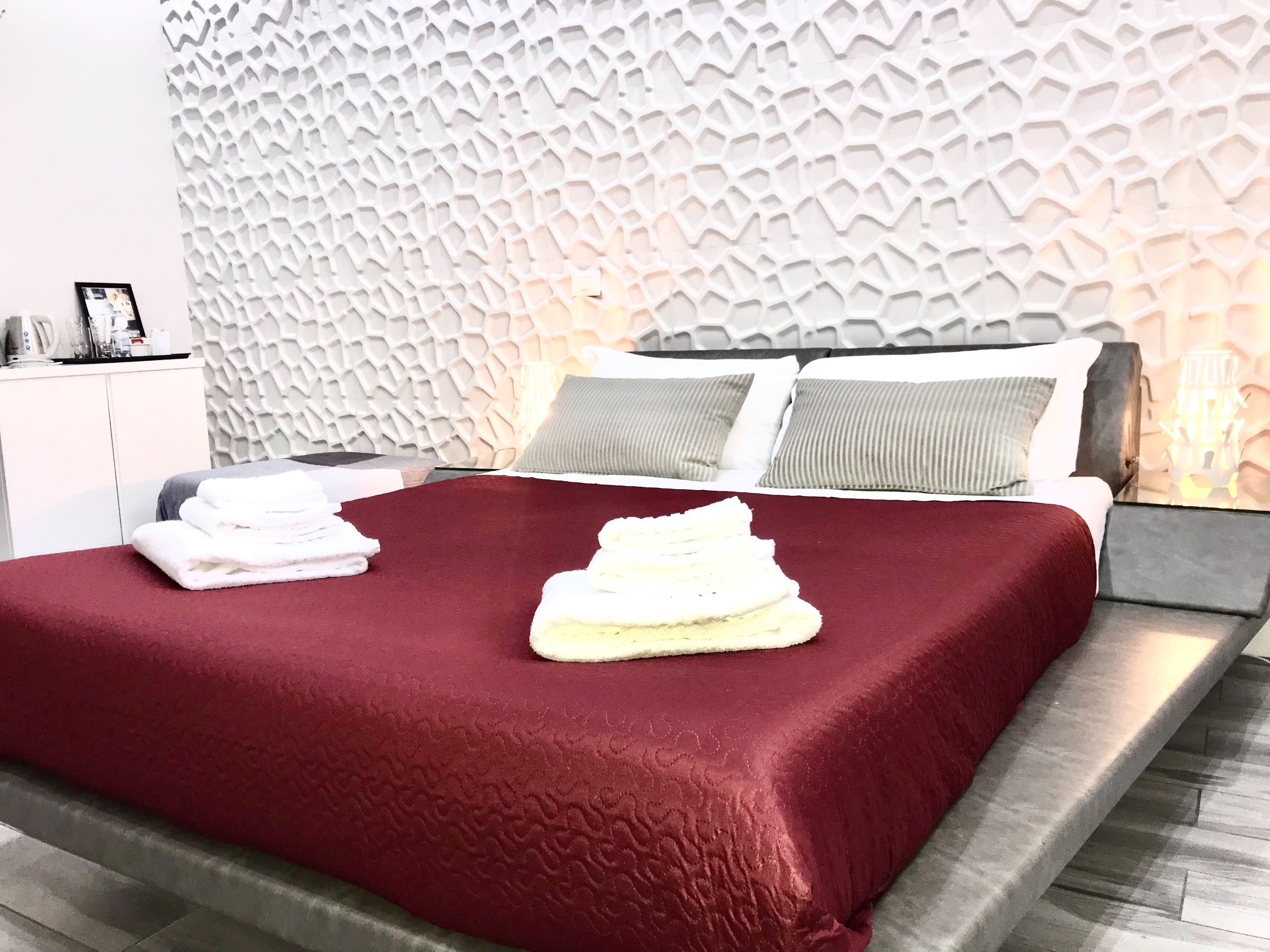 Quarto de hotel moderno com parede estampada em padrão geométrico, cama bem arrumada com colcha vermelha e uma pilha de toalhas ao lado.
