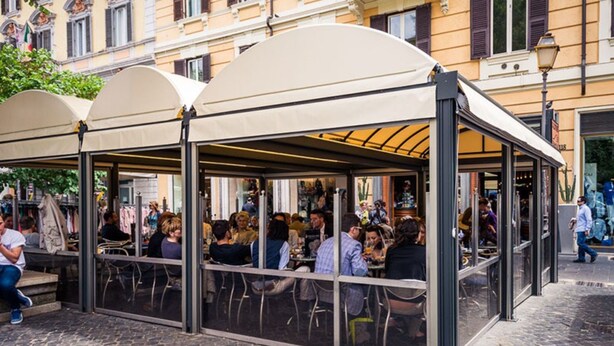 A covered outdoor seating area with tables and chairs, surrounded by a glass enclosure, in an urban setting with buildings in the background. - Domus Tacito Luxury (Rome)