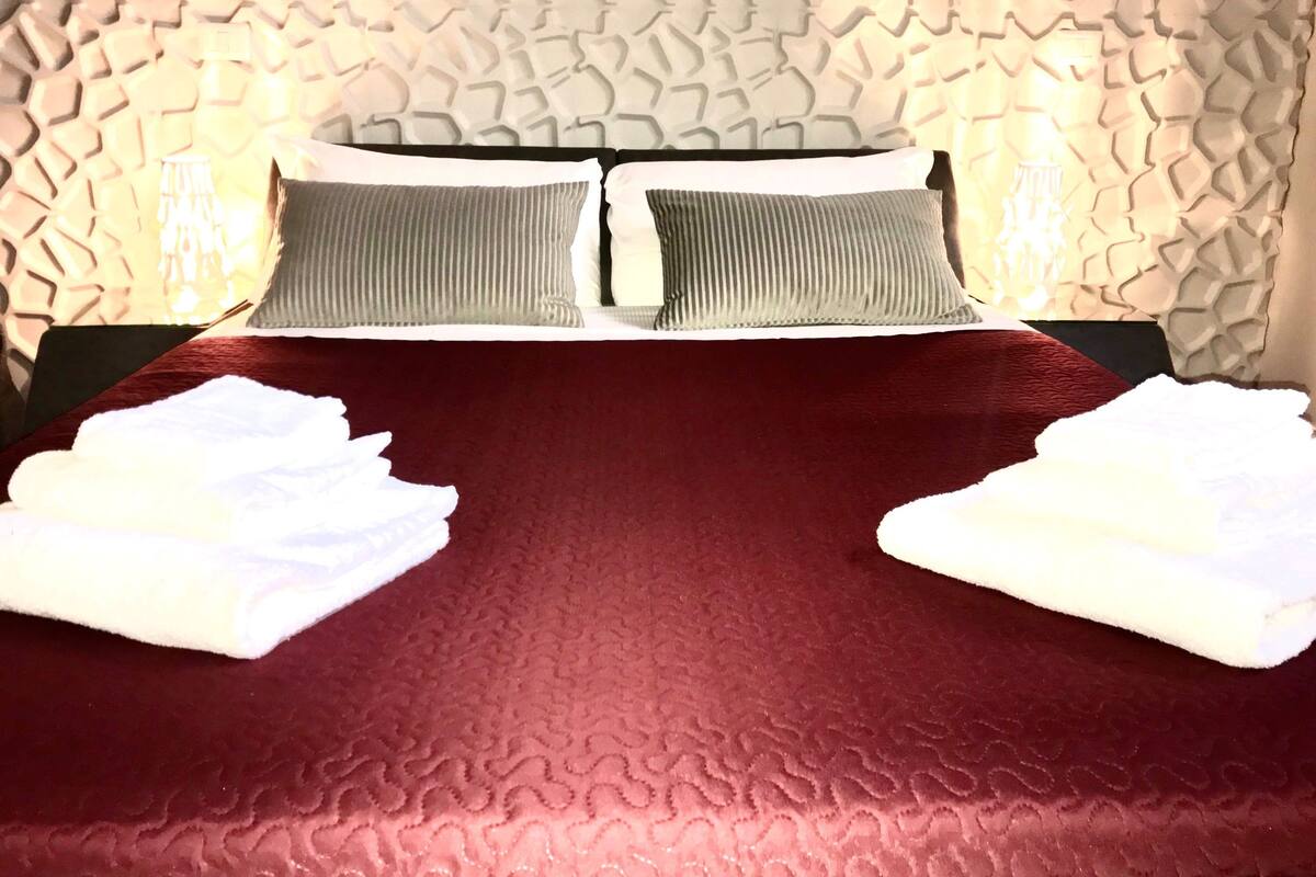 Una cama con una colcha roja, dos almohadas y dos toallas blancas dobladas.