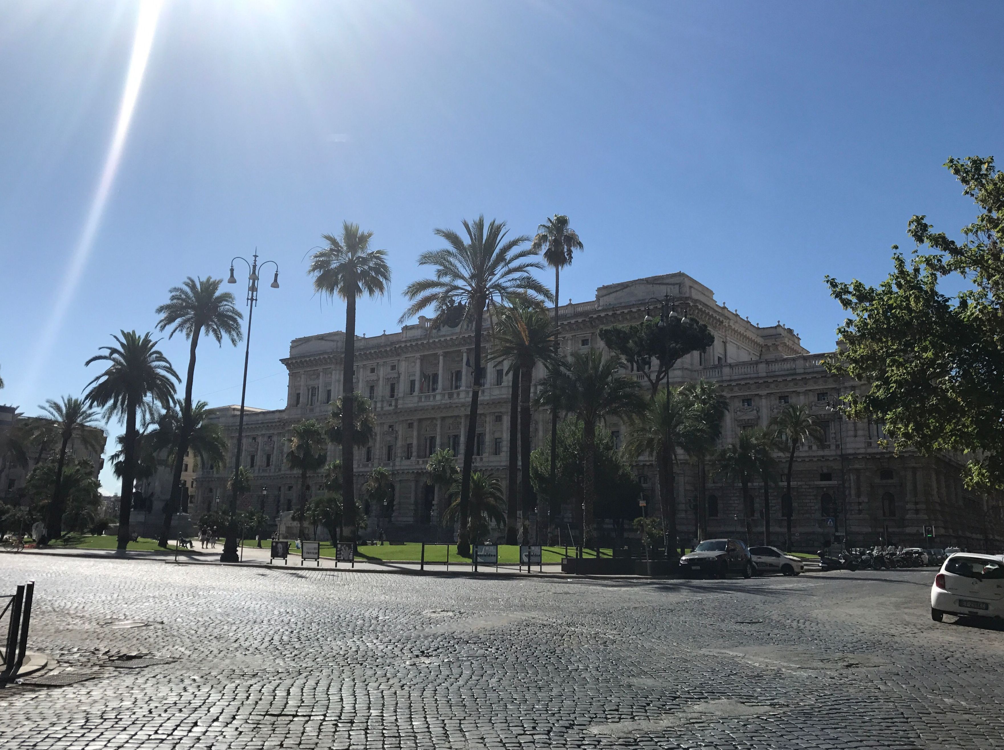 A large historic building with a symmetrical facade, surrounded by palm trees and a cobblestone plaza.