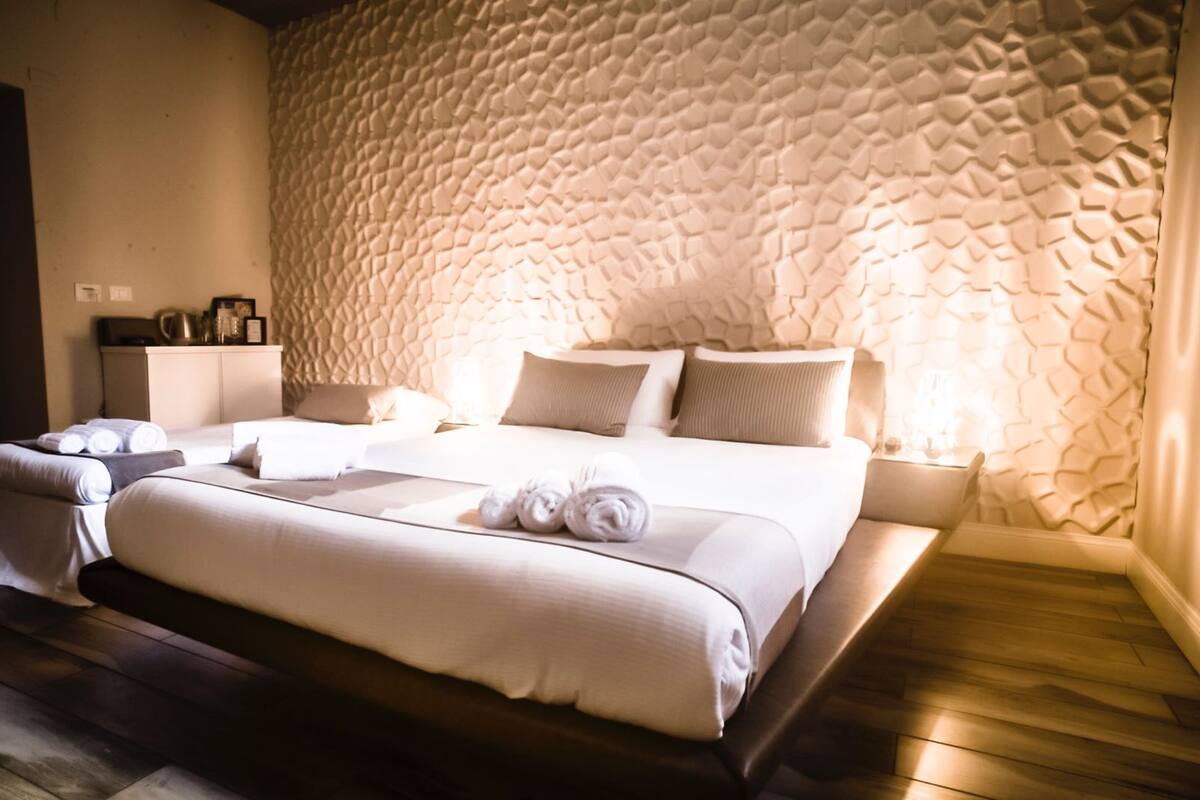 Habitación de hotel con pared texturizada, cama con ropa blanca, mesita de noche con lámpara y piso de madera.