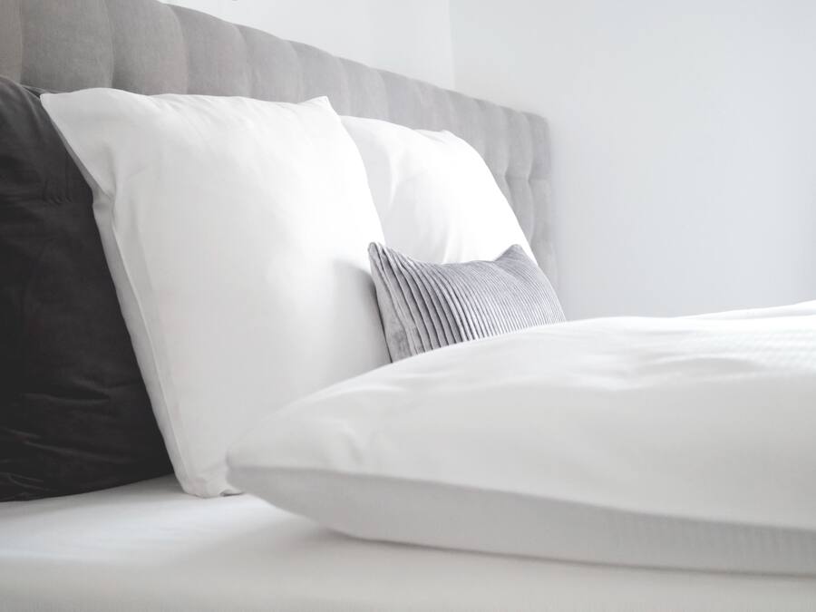 Una cama bien tendida con ropa de cama blanca y gris, un cabecero tapizado en gris y una almohada gris oscuro.