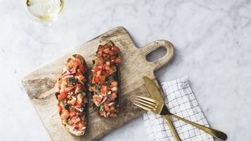 Two pieces of bruschetta on a wooden board with a fork and knife on a checkered napkin.