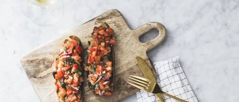 Dos bruschettas sobre una tabla de madera con un tenedor y un cuchillo sobre un mantel a cuadros.