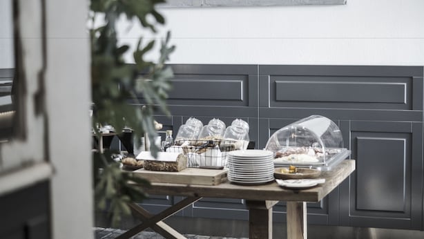 Una mesa de buffet con alimentos y cristalería, frente a una pared gris con paneles.