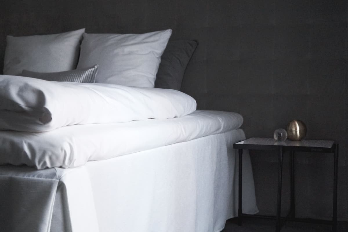 A neatly made bed with white linens and pillows, a small bedside table with two spherical objects, and a dark wall in the background.