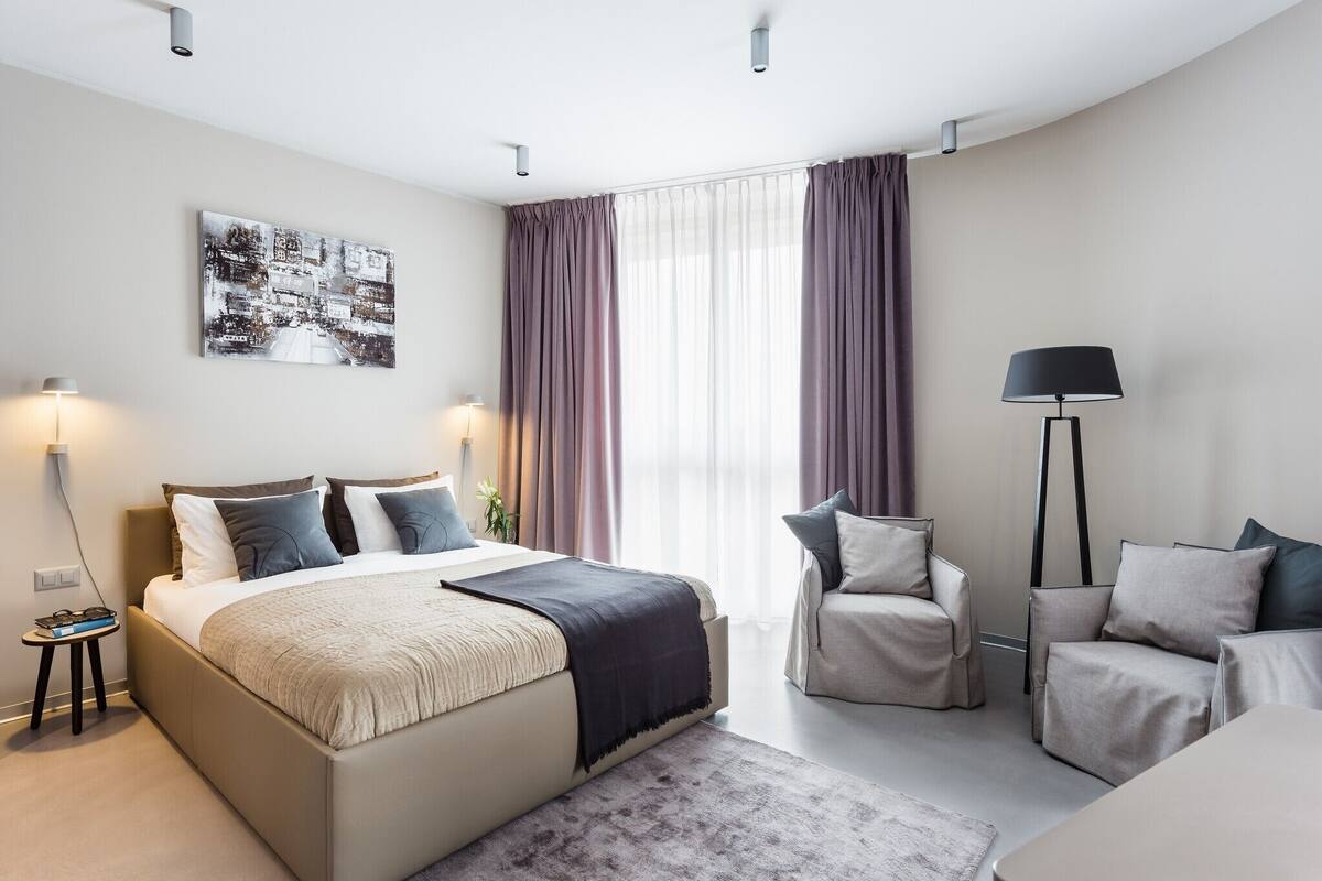 A modern hotel room with a large bed, two armchairs, a floor lamp, and a side table.