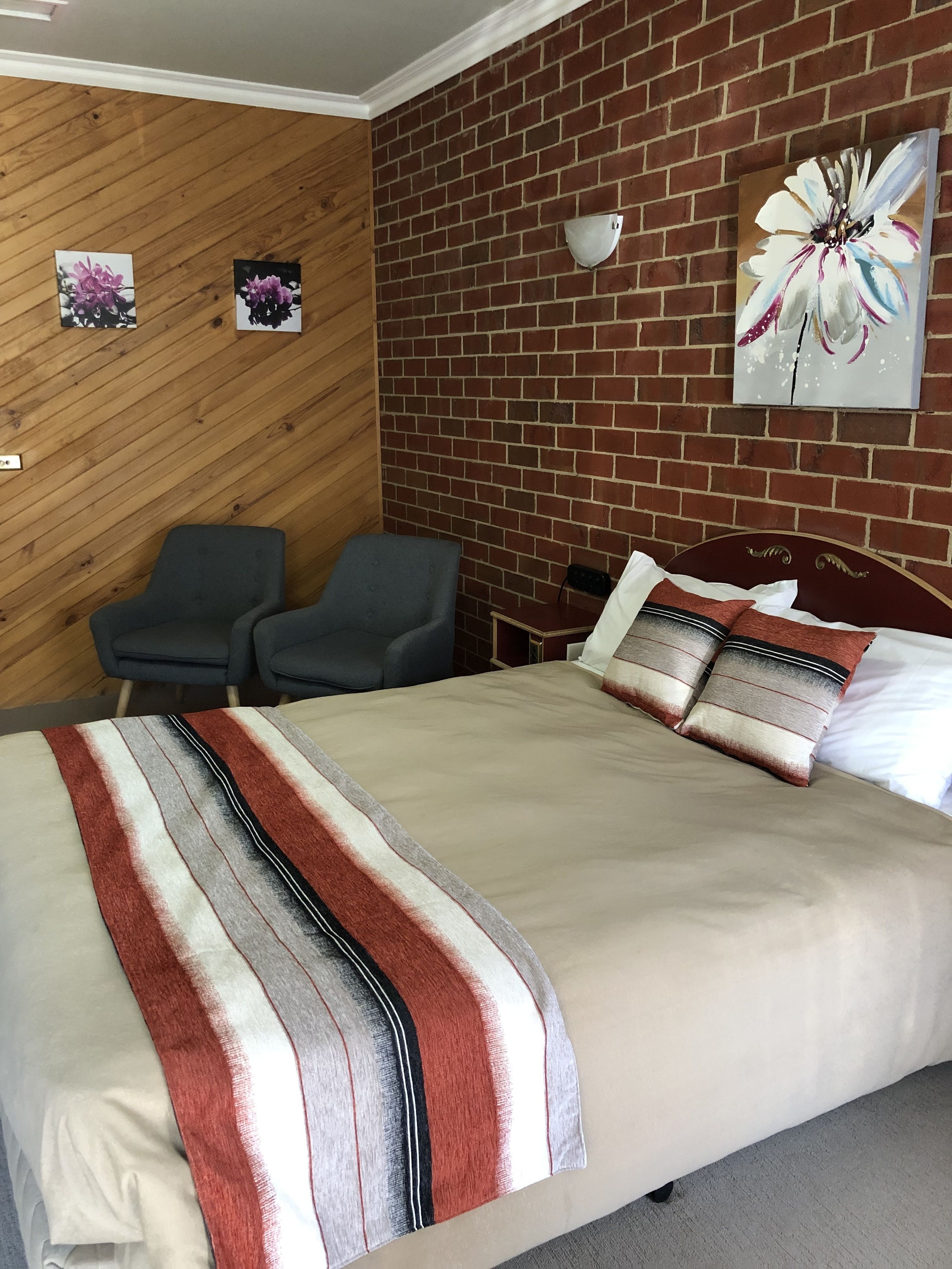 A hotel room with a large bed, two armchairs, a wooden wall, and a brick wall with a flower painting.