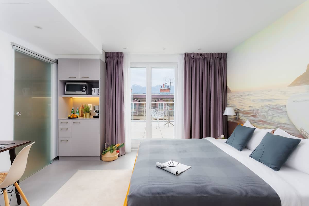 A modern hotel room with a bed, a small kitchenette, and a balcony with a view of the city.