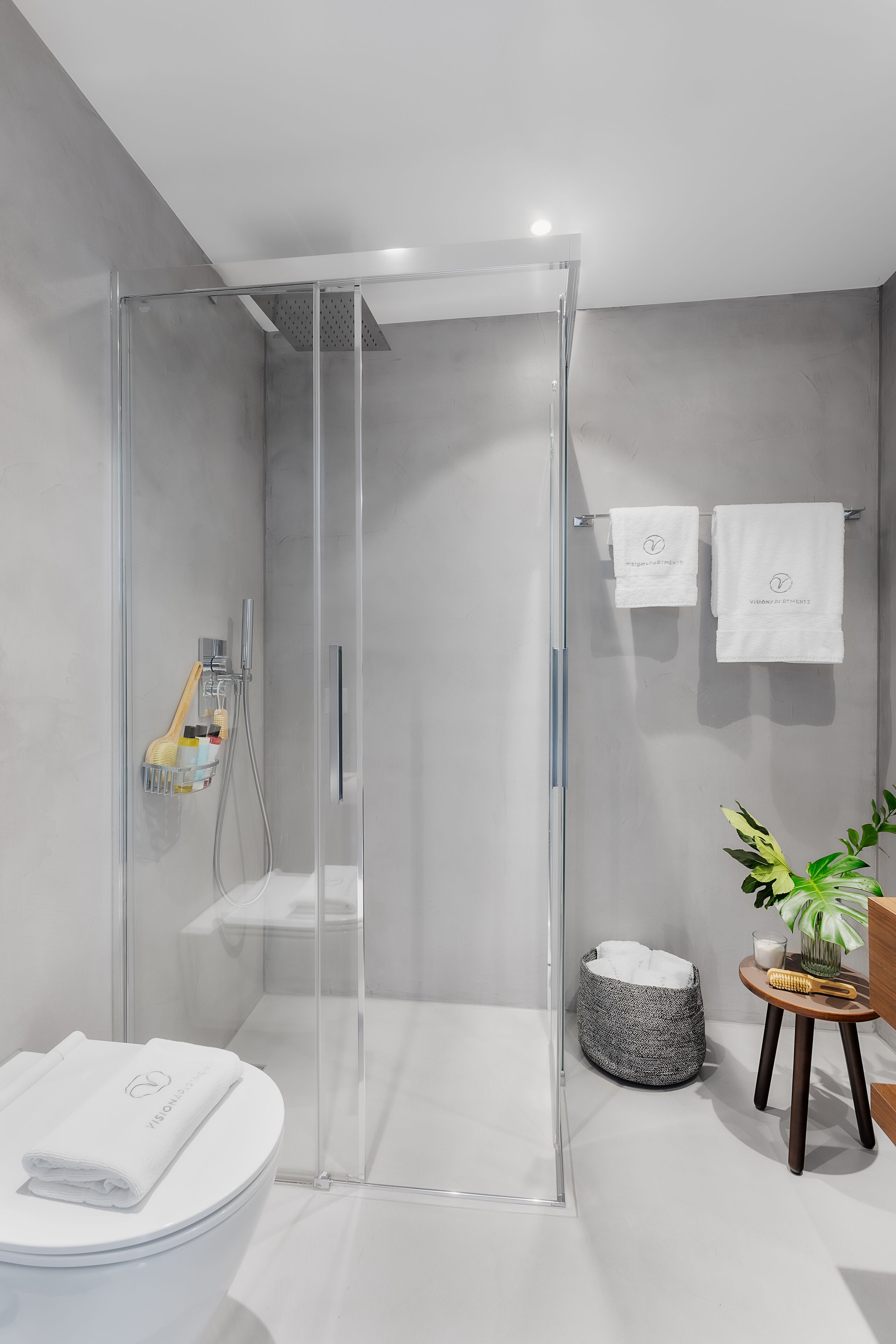 Een moderne badkamer met een douchecabine van glas, een toilet met een handdoek, een klein rond tafeltje met een plant en een grijze badmat.