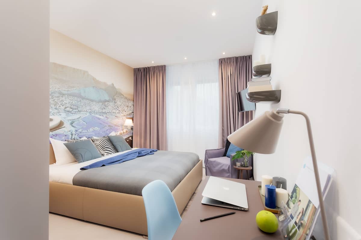 A modern hotel room with a bed, a desk with a laptop, a lamp, and a wall-mounted shelf with books and a plant.