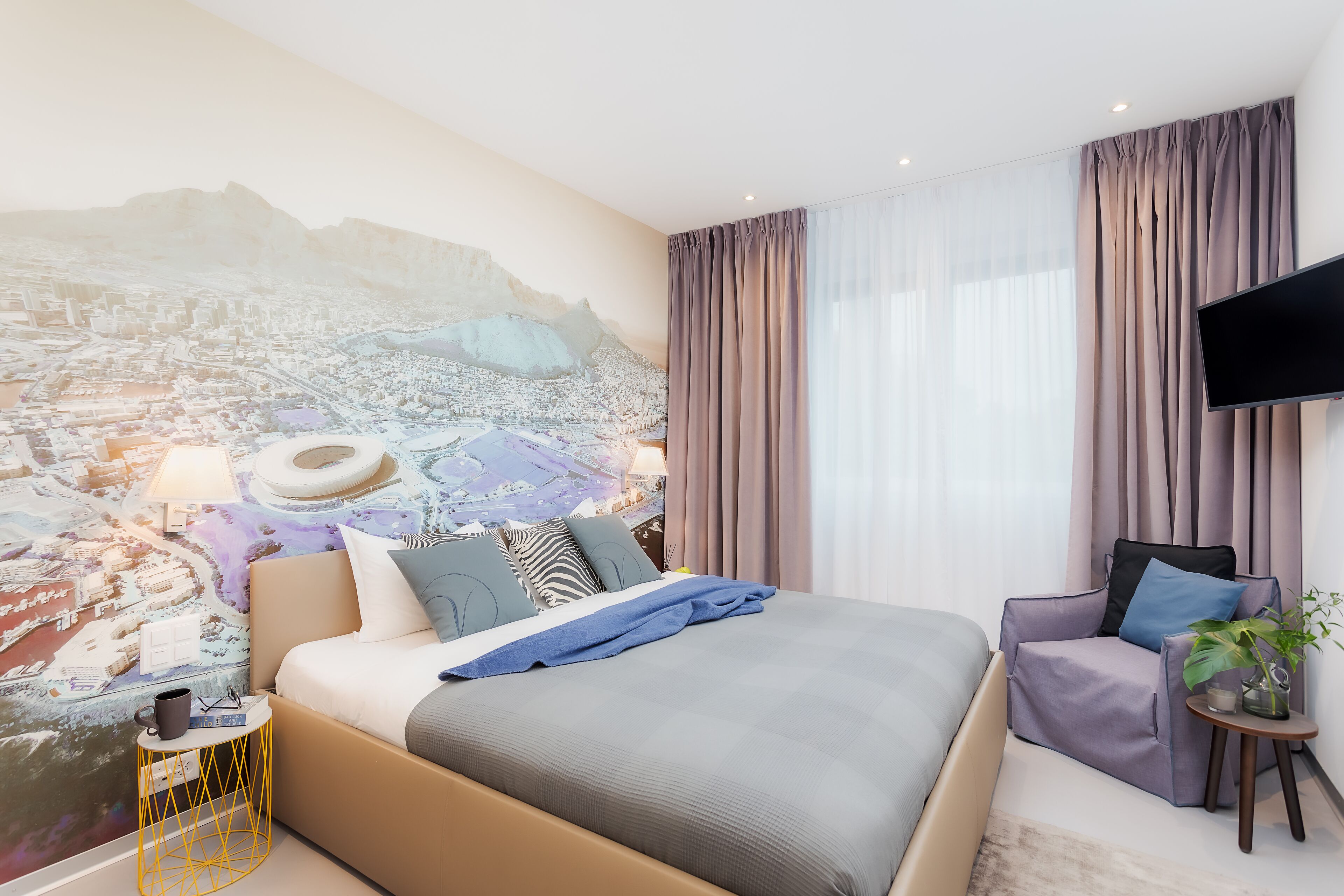 A modern hotel room with a large bed, a chair, a small table, and a wall-mounted TV.