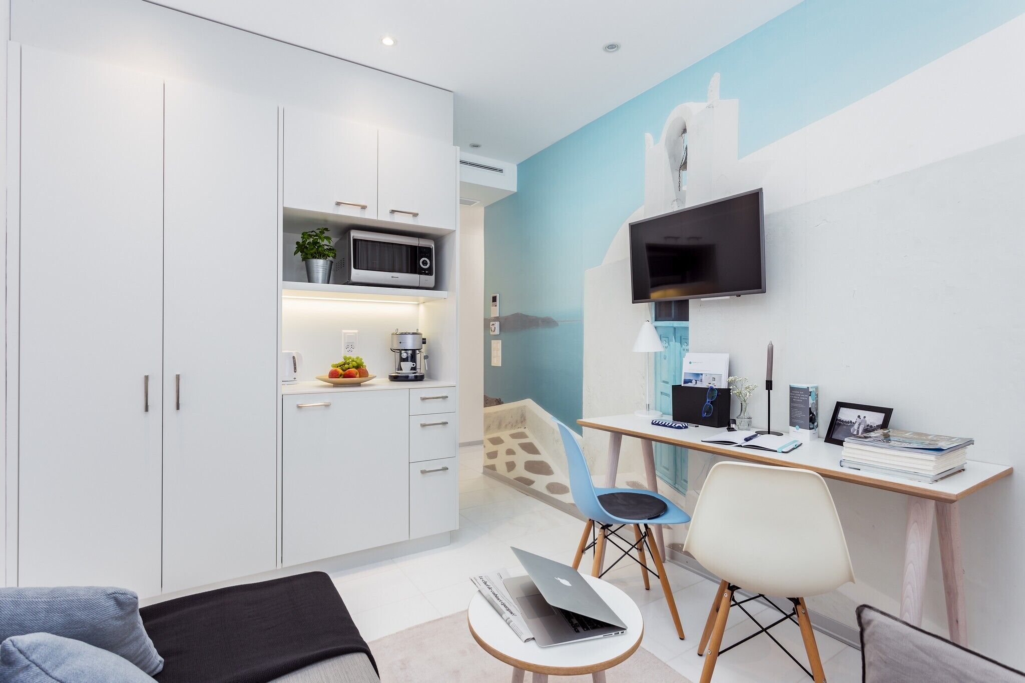 Una cocina moderna con armarios blancos, microondas y cafetera. Hay un escritorio con computadora, silla y libros. Un televisor está montado en la pared.