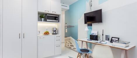 Una cocina moderna con armarios blancos, microondas y cafetera. Hay un escritorio con computadora, silla y libros. Un televisor está montado en la pared.