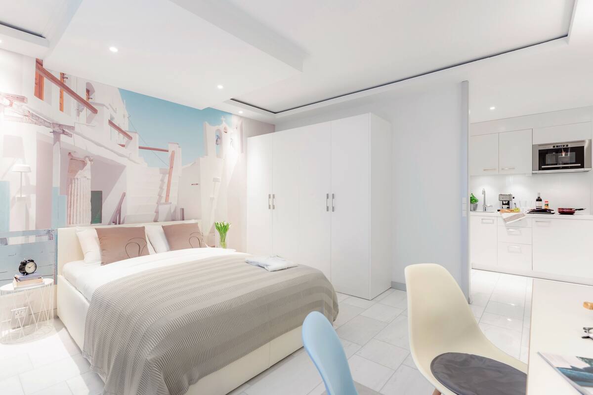 A modern bedroom with a large bed, a desk, and a kitchenette.