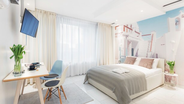 A modern hotel room with a bed, a desk with a chair, a TV, and a window with curtains.