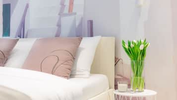 A neatly made bed with a white and beige color scheme, a small white side table with a glass vase containing green leaves, and a white wall with a partial view of a painting.