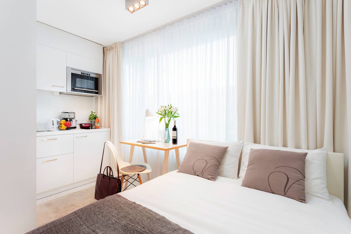 A modern hotel room with a bed, a desk, and a kitchenette.