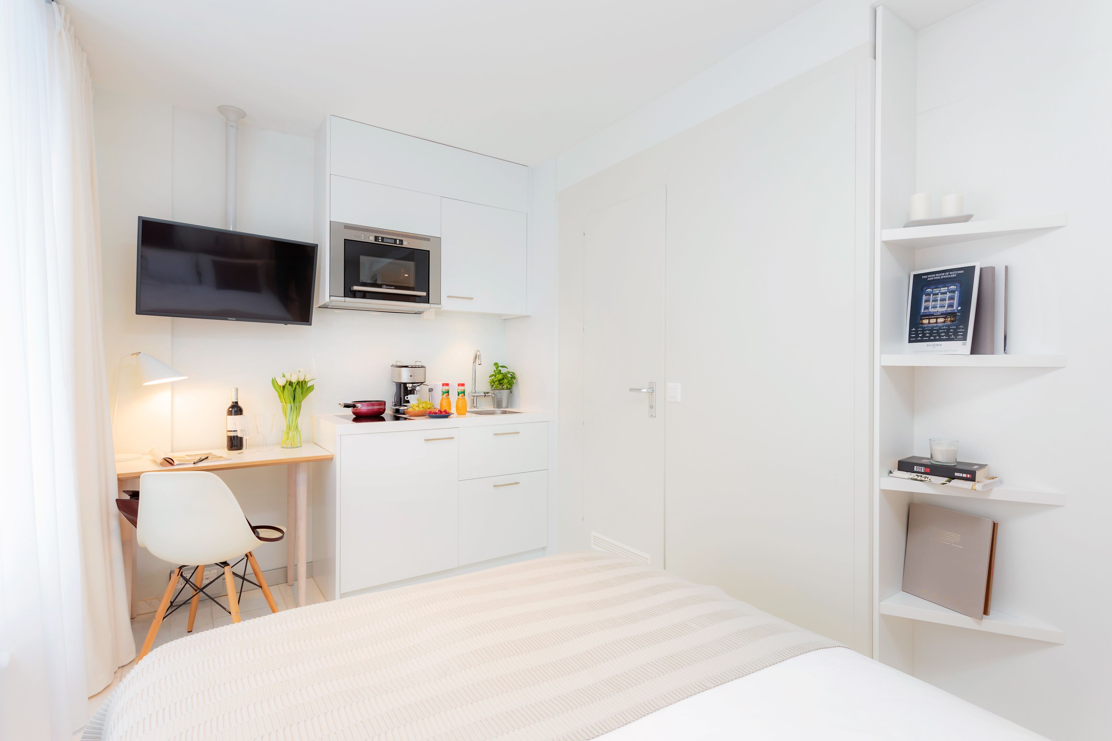 A modern hotel room with a bed, a desk with a chair, a TV, a microwave, and a bookshelf.