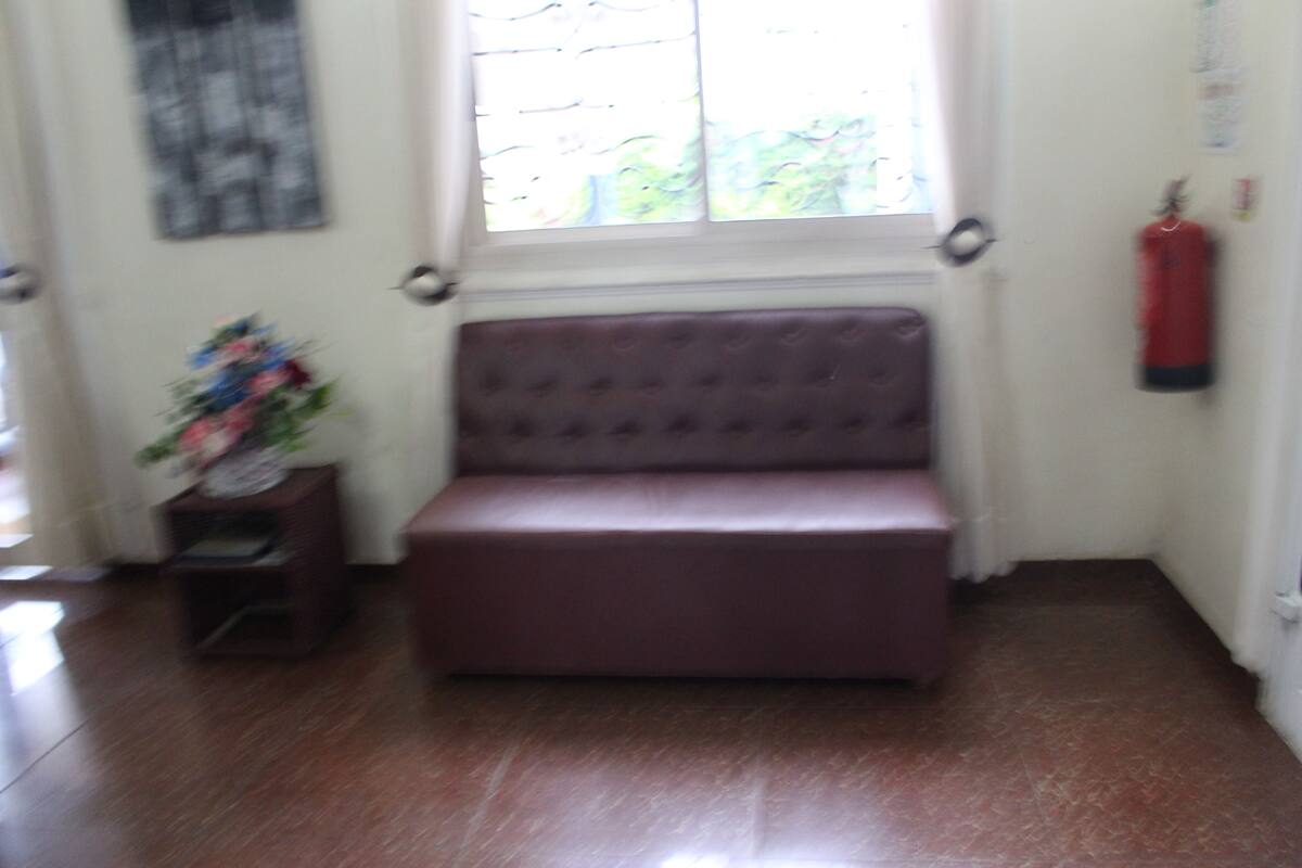 A room with a brown cushioned bench, a small wooden side table with a floral arrangement, a fire extinguisher on the wall, and a window with blinds.