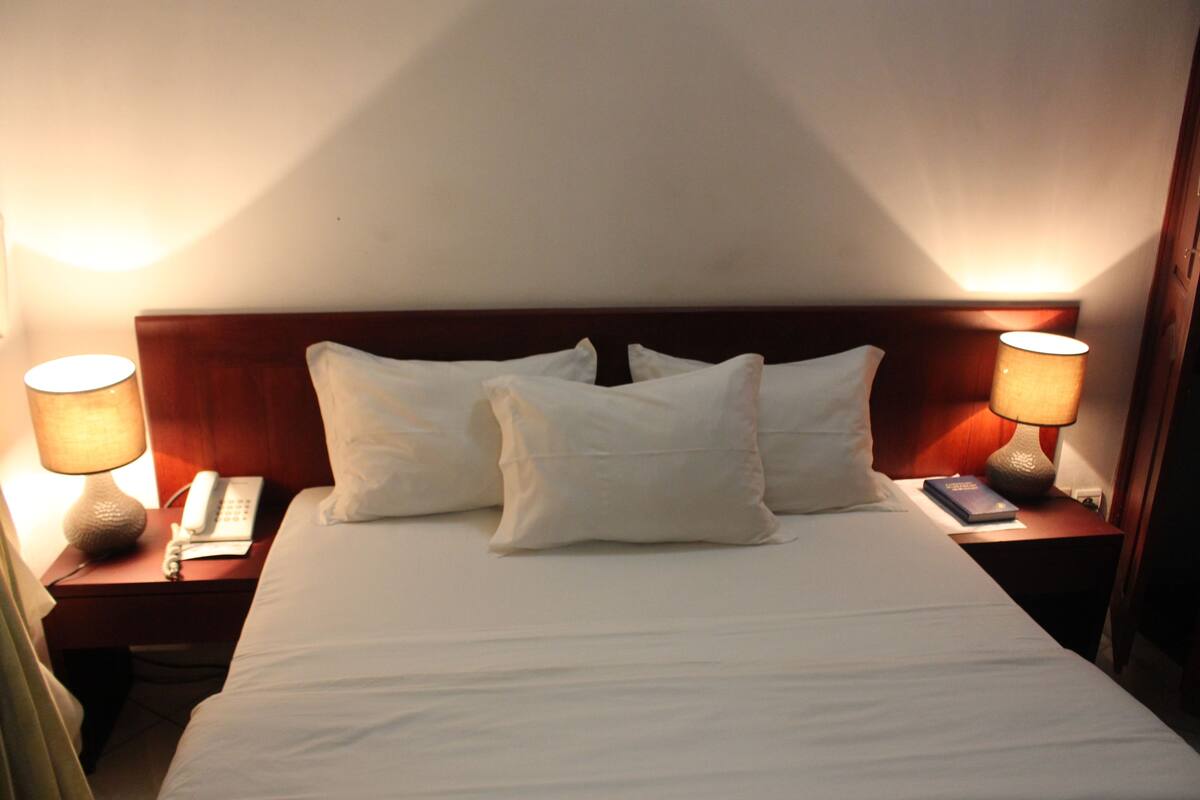 A hotel room with a neatly made bed, two bedside lamps, a telephone, and a book on the nightstand.