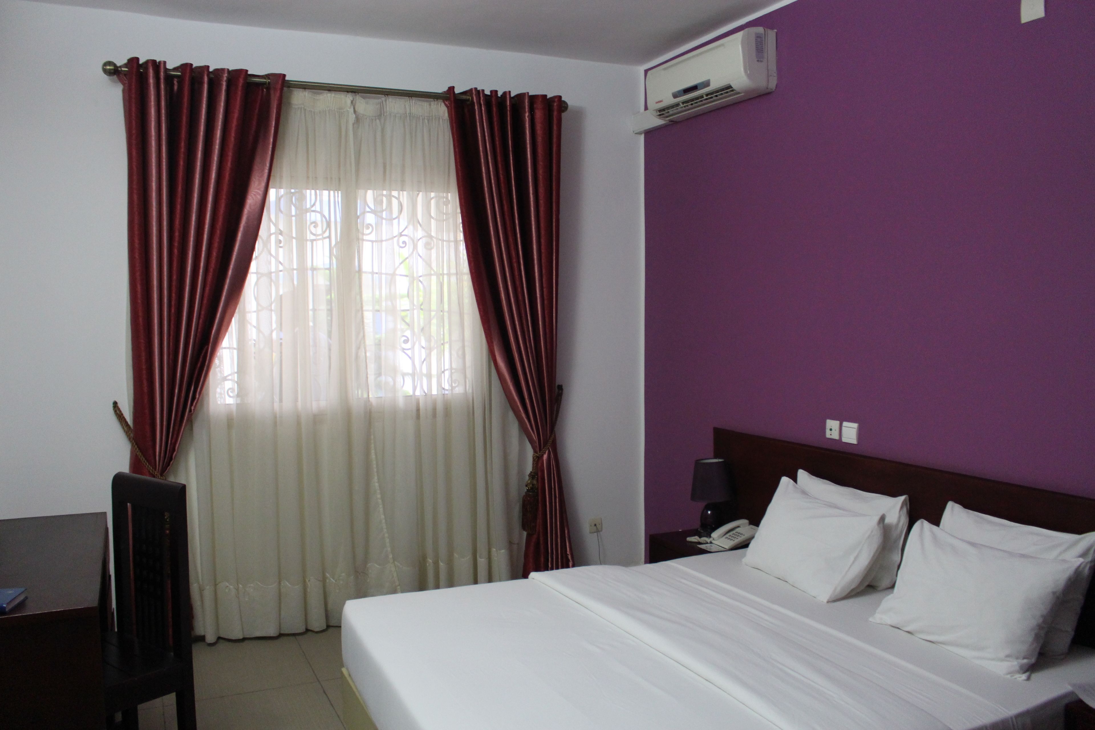 A hotel room with a bed, curtains, a desk, and an air conditioning unit.