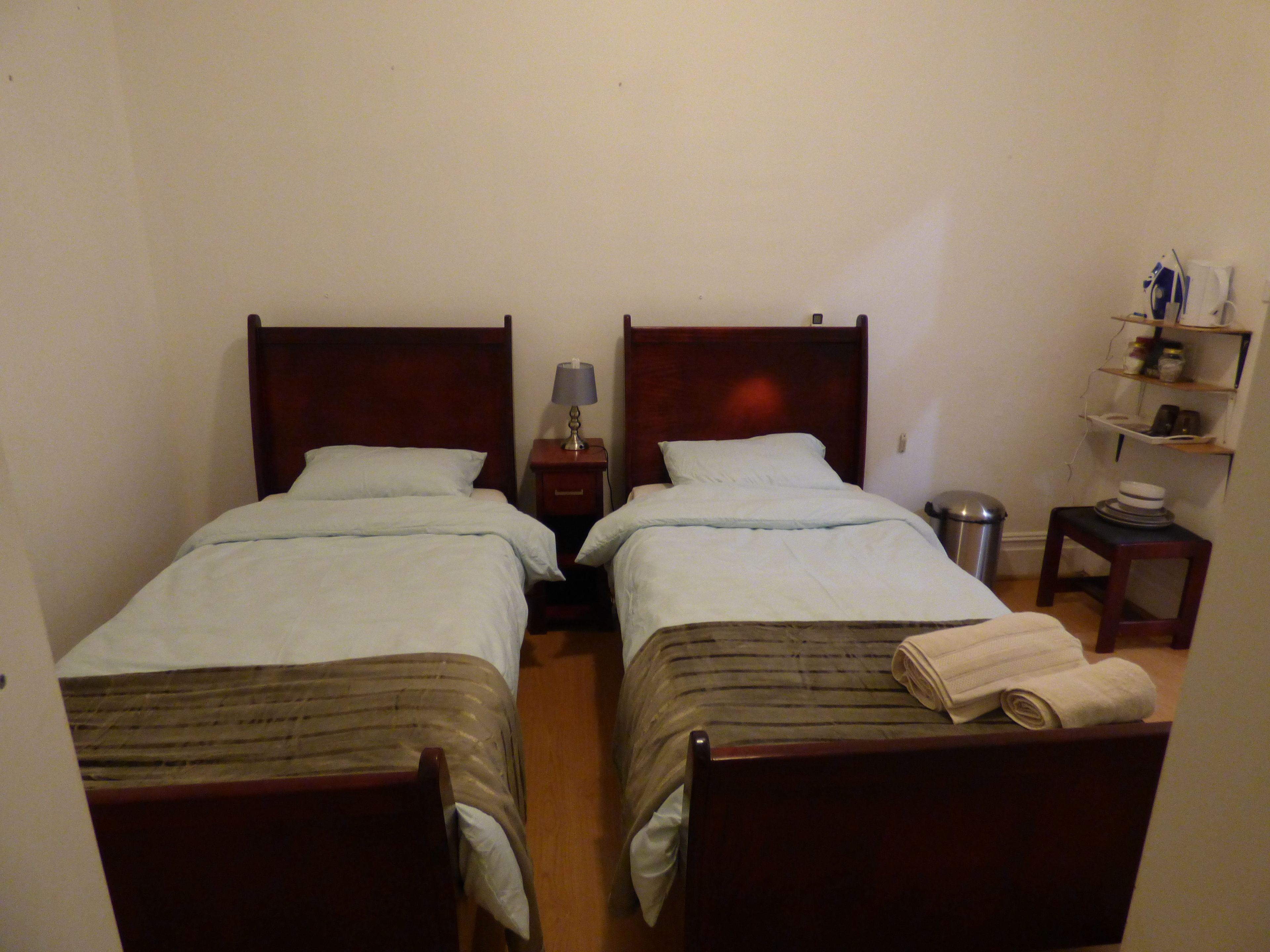Une chambre avec deux lits simples, une table de chevet avec une lampe, une petite poubelle et une étagère contenant divers objets.