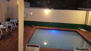 A swimming pool with a blue light, surrounded by a brick patio and white chairs. - Daisy Lodge (Durban)