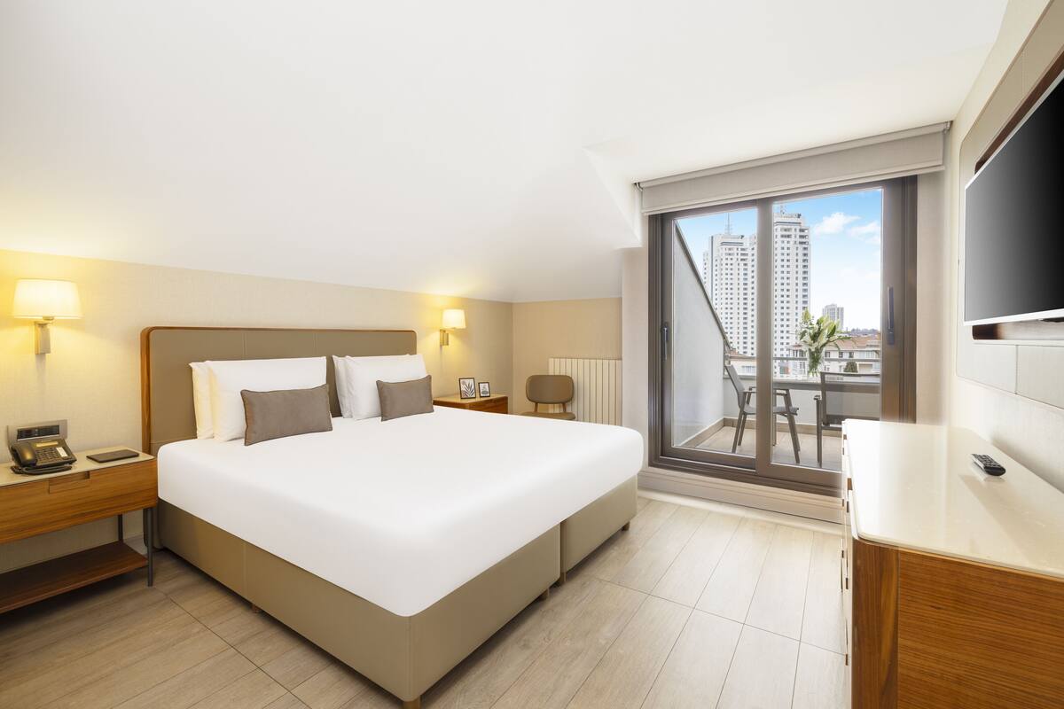 Hotellihuone, jossa on suuri sänky, yöpöydällä lamput, kirjoituspöytä puhelimineen ja suuri ikkuna, josta avautuu näkymä kaupunkiin.